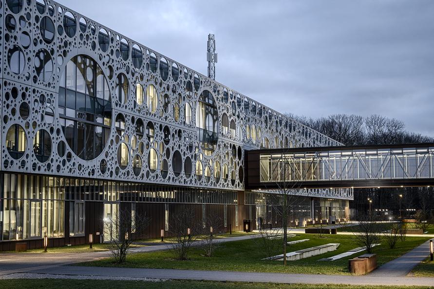 Modern building with a perforated metal facade, illuminated at dusk, surrounded by a landscaped area.