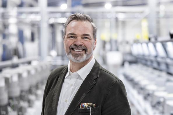 Portrait of a smiling man in a factory, standing in front of industrial machines with spools of thread.