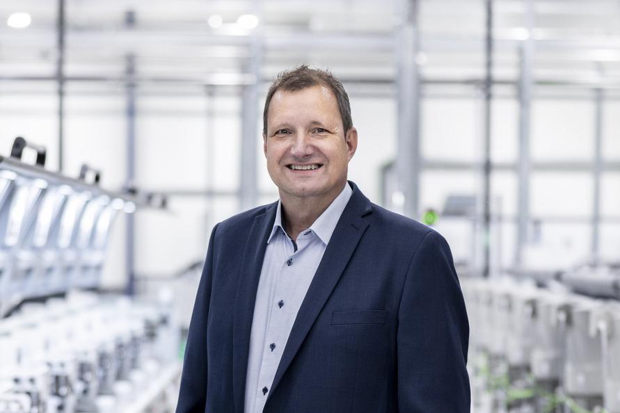 A man in a suit stands confidently in a modern manufacturing facility surrounded by machinery.