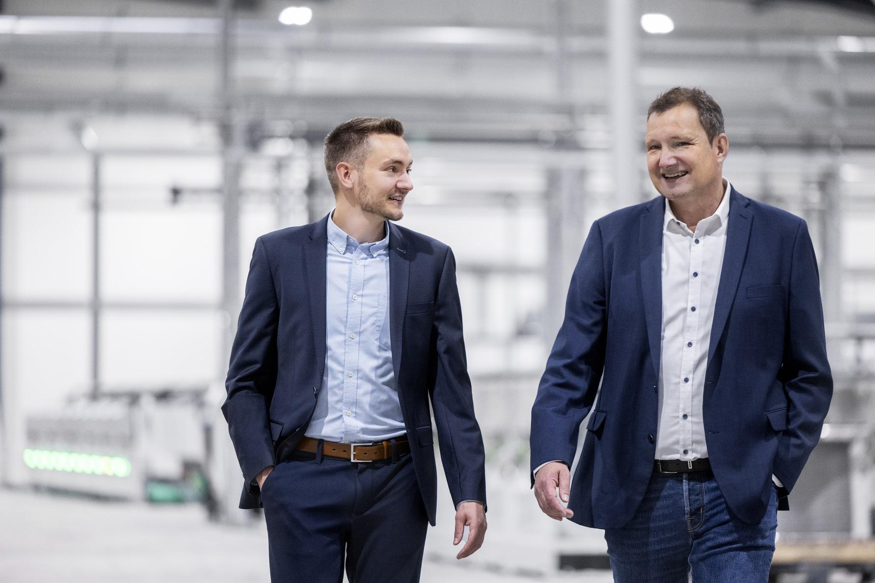 Two men in business suits walk together through a modern industrial space, engaged in conversation and smiling.