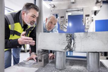 Two men observe a metal specimen being tested for pressure in a laboratory setting, examining the results closely.