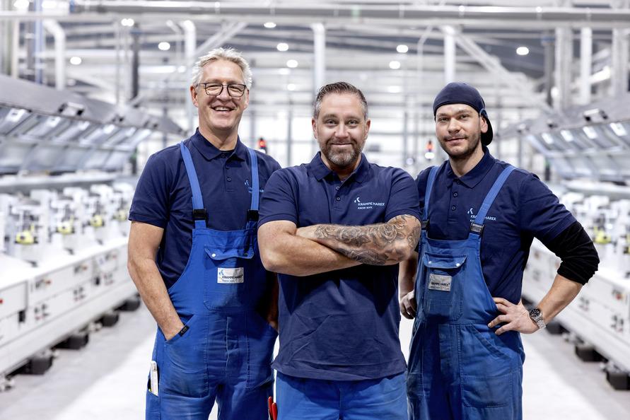 Three workers in blue overalls and dark shirts stand confidently in a factory with machinery in the background.